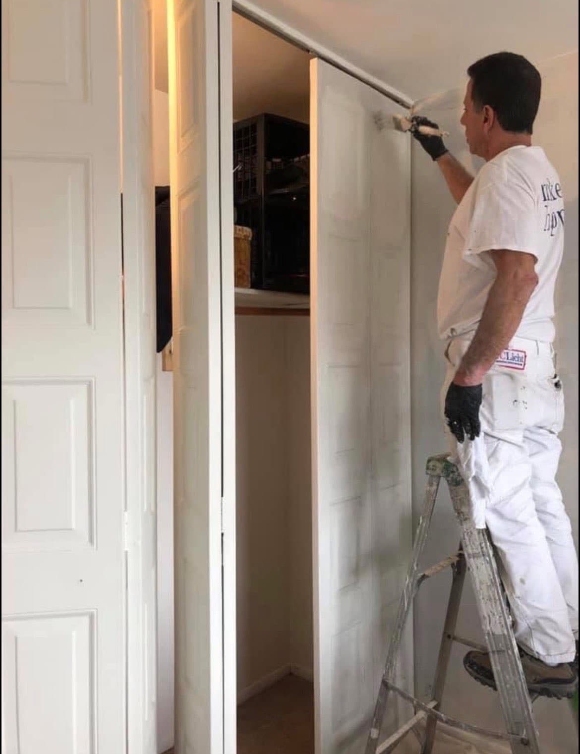 A professional painter on a ladder paints white bifold closet doors with a brush.